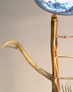 A close-up of this sculpture shows the upper left quarter of the art piece. The madrone wood of the ladder body is gold leaf on the outside and heartwood inside. A gold-leafed snake carved into the ladder cross back and forth to form the rungs that rising up to the head. Here a snake head appears as an eye, like the calmly lidded Sipa Barda's. Vibrant snake tongue extends across out of frame. The wooden ladder culminates in a feline shaped ear. The ear point holds up a partially visible convex cosmic egg in shimmering silver leaf, and blues. at the lower right side, partially visible at the center of the ladder body is a delicate brass spiral. To the left is a pronghorn antler reaching upward as its right arm. In its palm is held a moonstone cabochon.