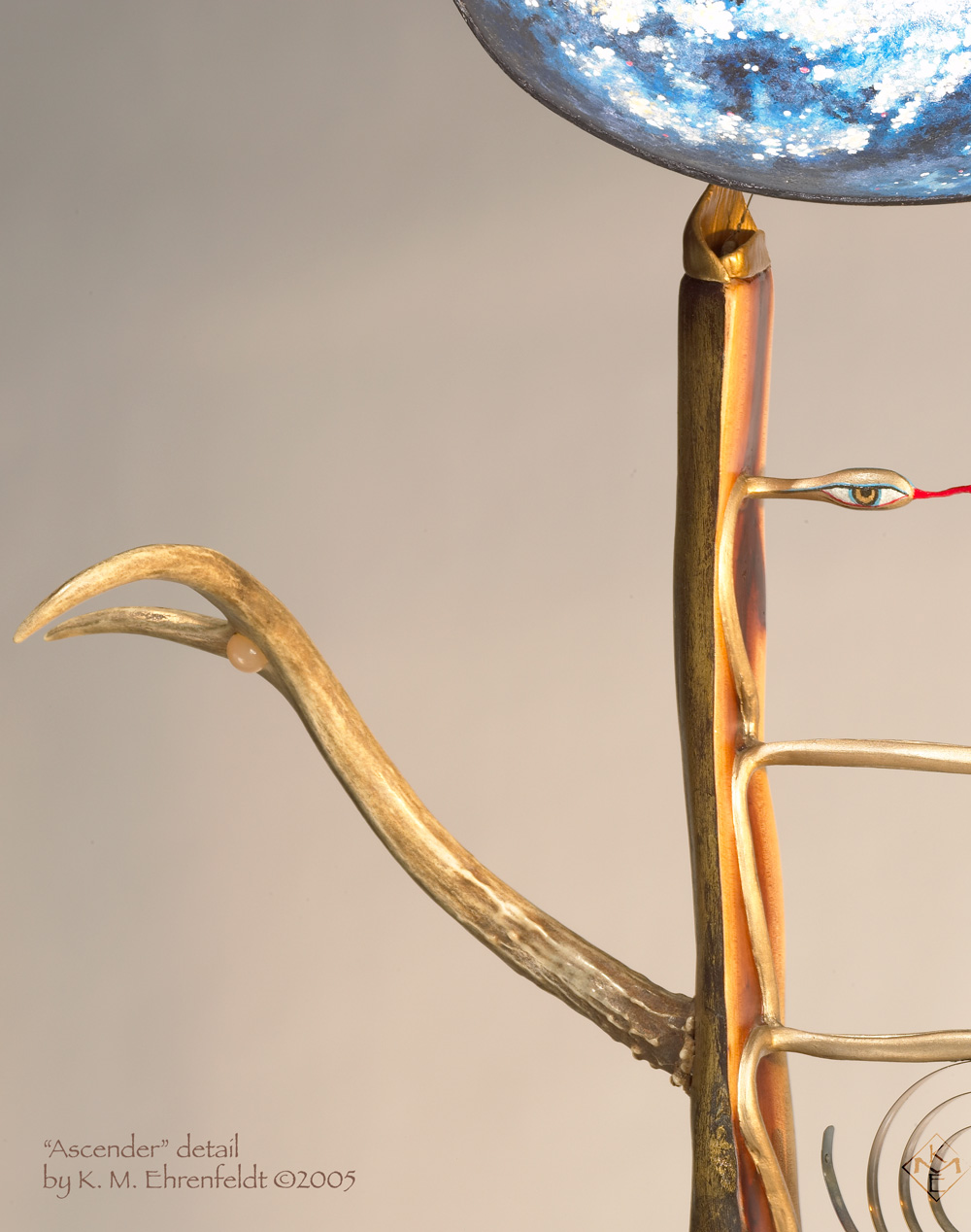 A close-up of this sculpture shows the upper left quarter of the art piece. The madrone wood of the ladder body is gold leaf on the outside and heartwood inside. A gold-leafed snake carved into the ladder cross back and forth to form the rungs that rising up to the head. Here a snake head appears as an eye, like the calmly lidded Sipa Barda's. Vibrant snake tongue extends across out of frame. The wooden ladder culminates in a feline shaped ear. The ear point holds up a partially visible convex cosmic egg in shimmering silver leaf, and blues. at the lower right side, partially visible at the center of the ladder body is a delicate brass spiral. To the left is a pronghorn antler reaching upward as its right arm. In its palm is held a moonstone cabochon.