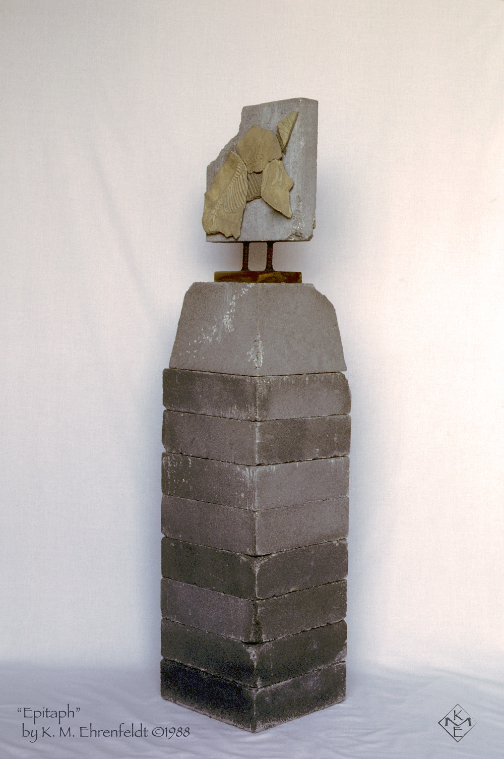 Rising up on a stack of eight cement bricks, each worn at the edges, sits a cement foundation block partly eroded at the top edge where the wooden insert extends up. From this wooden anchor, two rebar rods about four inches tall hold a cement paving stone up vertically. The paving stone has the upper left corner broken off. On this back facing paving stone are large ceramic shards pieced together and featuring the etchings of a horse's skeleton. The shard pieces together suggest the profile of a horse's head.