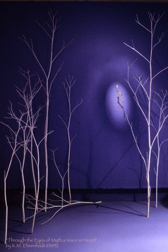 Suspended in the blue-violet room with a soft spotlight illumination is a twisting gold-leafed covered stick that appears to have decoration. The light illuminating it casts an egg-shape on the wall with additional radiating bands of light beyond. The piece is suspended from a delicate white tree, and to the left across this space are more white trees.
