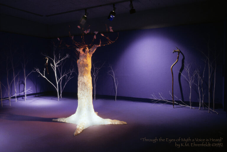 The blue-violet walls and floor of this room have a lightly wooded forest of white trees in the background. In the center of the room is a life-sized copper wire sculpture of a woman's form as a tree with four roots stretching in four directions. Her head and arms rising upward to form branches that reach outward and upward. Papery leaves dot the branches above. Her tree body is covered with a flakes of ivory rice-paper bark. She is lit from within at the base with a white light, and a magenta gel lighting illumines her from above. Leaning on the wall behind, on the right side, is a staff in the midst of the trees. In the far corner behind and to the left is a talking stick suspended in a white tree that is brightly lit. In a spot illumined on the left wall is a twisting gold-leafed covered stick that appears to have decoration. The light illuminating it casts an egg-shape on the wall with additional radiating bands of light beyond. In the middle ground are the broken column-shaped Core Sample sculptures, one laying on the ground parallel in view, the other standing vertically further back and to the right. These are softly lit to reveal stone-like surfaces of smooth gray, magenta, and blue transparent washes. White handprints and symbols are lightly etched onto the surfaces.