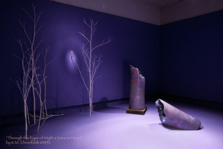 The blue-violet walls and floor of this room has a lightly wooded forest of white trees on the left side. In a spot illumined on the left wall is a twisting gold-leafed covered stick that appears to have decoration. The light illuminating it casts an egg-shape on the wall with additional radiating bands of light beyond. In the middle ground are the broken column-shaped Core Sample sculptures, one laying on the ground parallel in view, the other standing vertically further back and to the right. These are softly lit to reveal stone-like surfaces of smooth gray, magenta, and blue transparent washes. White handprints and symbols are lightly etched onto the surfaces.