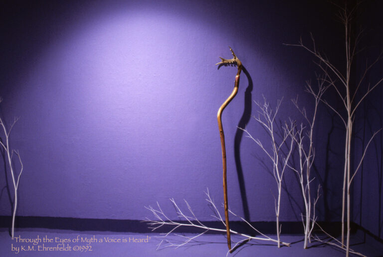 Leaning on the wall in a blue-violet room with delicate white trees creating a wooded atmosphere, the staff stands nearly 6 and 1/2 feet tall. The curved redwood branch is topped by a 4-point deer antler with eleven small brass bells attached to it. The redwood, freed of bark, is smooth with a sheen to it and there's a red-violet transparent streak flowing down it. A reddened band of coiled copper wraps around the top of the redwood staff a few inches below the base of the antler and again a few inches above the pointed end of the wood. The staff casts a curving shadow on teh wall behind.