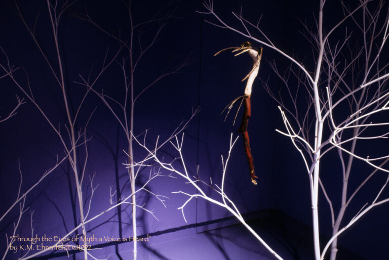 Suspended in the blue-violet room with bright spotlight illumination is the curving madrone wood talking stick sculpture. It has the deep red-brown, smooth, skin-line bark on the lower half, and the creamy-colored heart wood visible in the upper half. The upper half serves as the body of the Storyteller bird's skeletal shape. The head is an actual bird skull with a long open-mouthed beak, it has a bronze-hued finish, with coiled copper at the neckline. At the curved shoulder of the stick, two bronze, bone-like, wing appendages reach outward. The body has been lightly wood burned to form delicate tattoo designs spiraling down the body from the neckline. Four bronze bone-like tail feathers jut out at the base of the body where the madrone bark begins. The lower half of the stick culminates in a naturally weathered break that is a reaching hand shape. White trees fill the background casting their shadows on the wall behind.