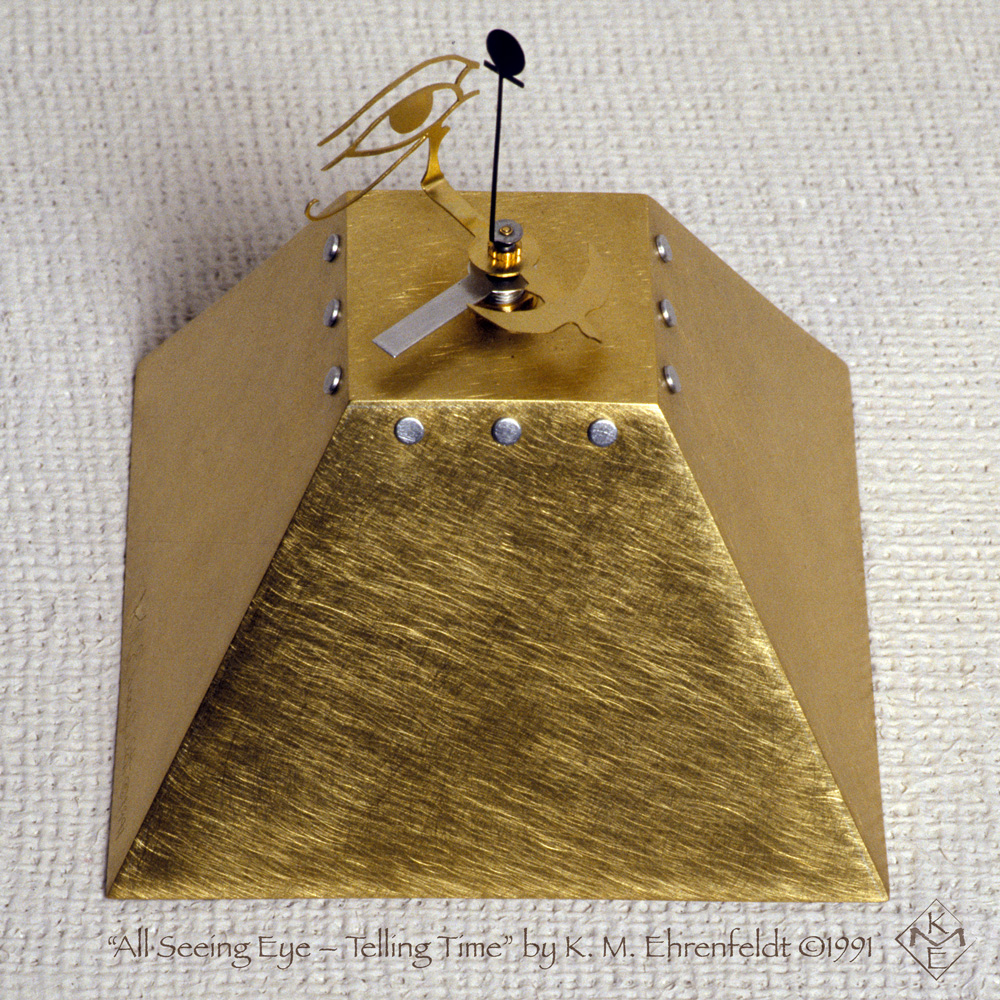 Looking down onto the top of the sculptural clock, the flat-topped, four-sided pyramid is topped with four sculptural forms that will move in clock rotation. The second hand rising straight up is a black Shen (eternity symbol of a disc standing on a horizontal rod). The minute hand is a brass hawk silhouette pointing outward parallel to the top of the pyramid, and opposite the minute hand representing watchfulness – a cutout brass Wedjet-eye is positioned vertically to be about the same height as the rod holding up the Shen. The hour hand is a flat aluminum plank parallel to the pyramid top. On each side of the pyramid, at the top edge are 3 aluminum discs marking the hours. The edges of the pyramid are seamless, providing clean lines to the structure itself. The brass surface has a brushed texture that spreads a textured light across its surface.