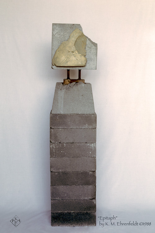 Rising up on a stack of eight cement bricks, each worn at the edges, sits a cement foundation block partly eroded at the top edge where the wooden insert extends up. From this wooden anchor, two rebar rods about four inches tall hold a cement paving stone up vertically. The paving stone has the upper right corner broken off. On this front facing paving stone is a large ceramic shard featuring a skeletal etching of an animal's leg bones.