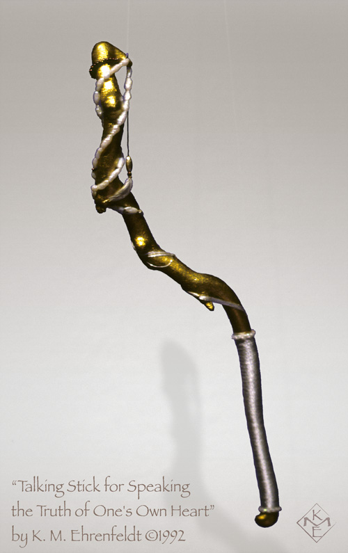 A twisting gold-leafed covered stick of madrone wood has a wrapped ivory-hued silky cord covering for one third of the length of the stick to serve as the handle. Two freshwater pearl strands wind loosely down in a twisted drape from the top to about a third of the way down the stick. Mahogany colored seed beads encircle the tip of the stick, with a silky thread dangling down a few inches with a silver bead and small white feather at the end.