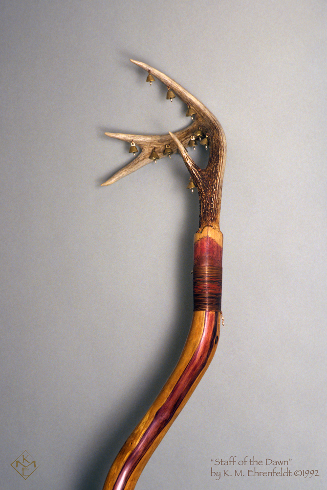 In this close up of the top of the curved redwood branch, topped by a 4-point deer antler with eleven small brass bells attached to it. The redwood, freed of bark, is smooth with a sheen to it and there's a red-violet transparent streak flowing down it. A reddened band of coiled copper wraps around the top of the redwood staff a few inches below the base of the antler and there are silver beads at the start and end of the coiled copper. a silver bead hangs beneath each of the bells.
