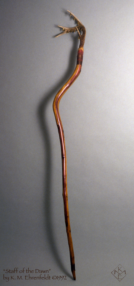 Standing nearly 6 and 1/2 feet tall, the curved redwood branch is topped by a 4-point deer antler with eleven small brass bells attached to it. The redwood, freed of bark, is smooth with a sheen to it and there's a red-violet transparent streak flowing down it. A reddened band of coiled copper wraps around the top of the redwood staff a few inches below the base of the antler and again a few inches above the pointed end of the wood.